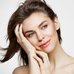 Beautiful naked young girl with perfect clean skin smiling touching hair over white wall. Facial treatment. Copy space.